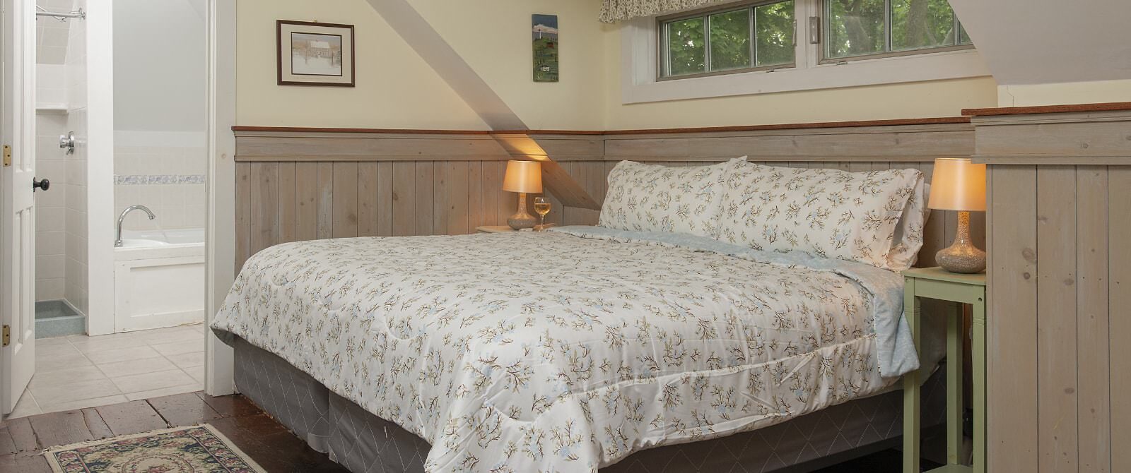 Bedroom with white walls, light wooden wainscoting, floral bedding, and view into bathroom