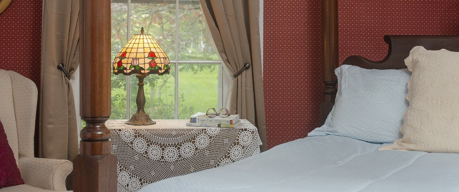 Close up view of four-poster dark wooden bed with light blue bedding, burgundy wallpaper, sitting area, and window with a view of the backyard