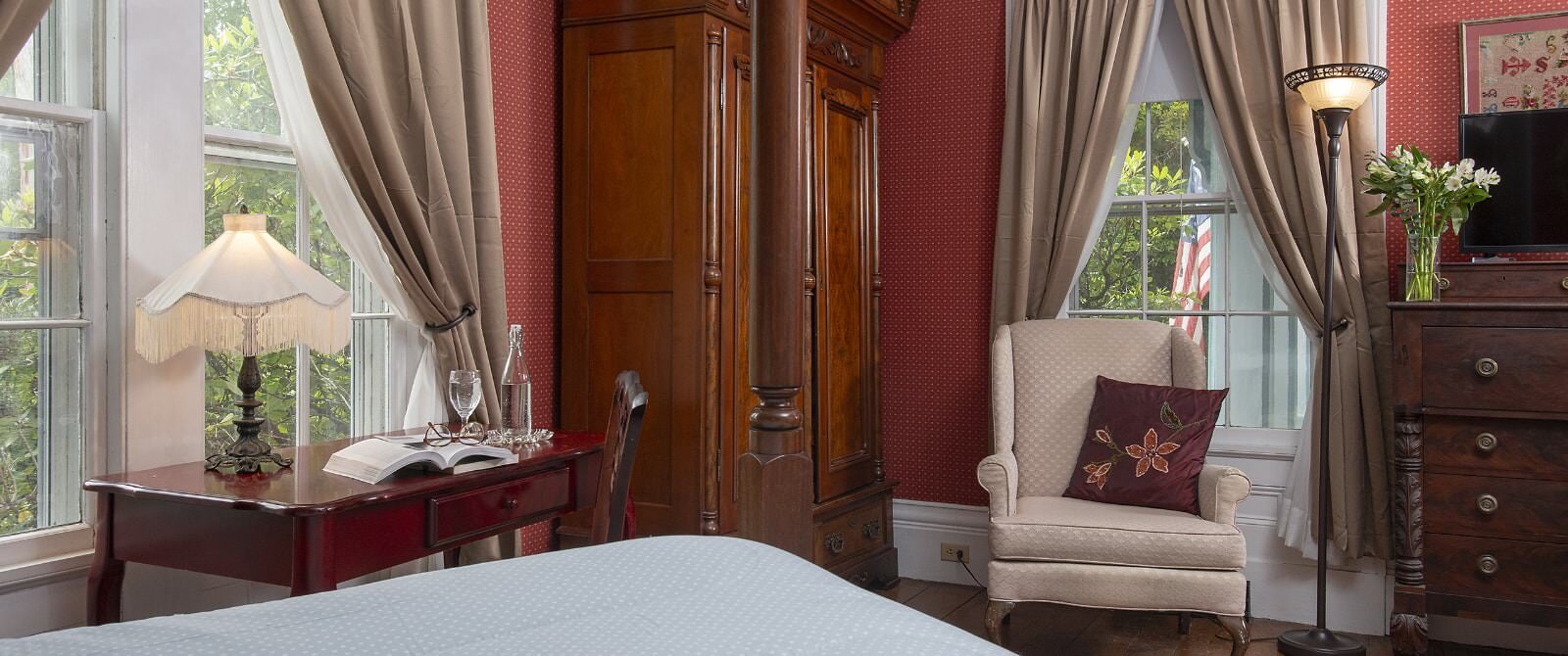 Bedroom with burgundy wallpaper, hardwood flooring, bed with light blue bedding, wooden desk, cream upholstered antique arm chair, and dark wooden hutch and dresser
