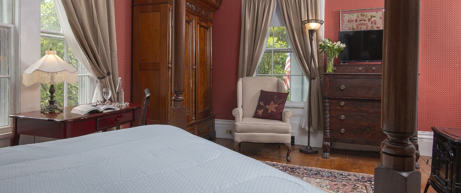 Bedroom with burgundy wallpaper, hardwood flooring, bed with light blue bedding, wooden desk, cream upholstered antique arm chair, and dark wooden hutch and dresser