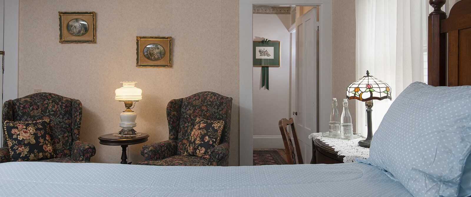 Bedroom with light peach wallpaper, antique wooden headboard, light blue bedding, sitting area, and antique wooden desk with chair