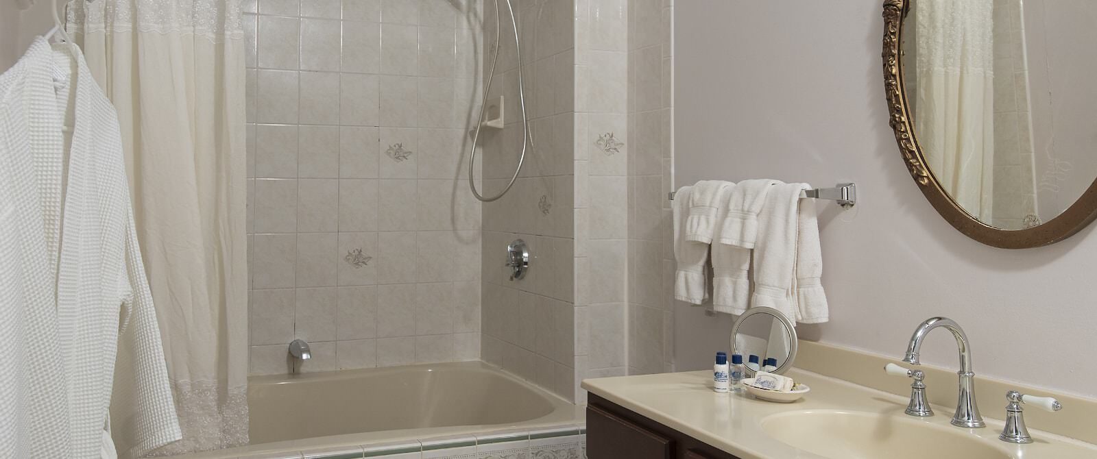 Bathroom with light walls, light tiled bathtub with shower, dark wooden vanity with cream counter, and oval framed mirror