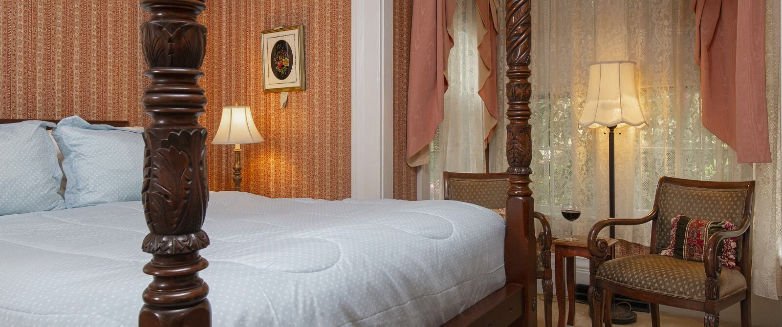 Bedroom with light colored striped wallpaper, carpeting, dark wooden four-poster bed, light blue bedding, and sitting area
