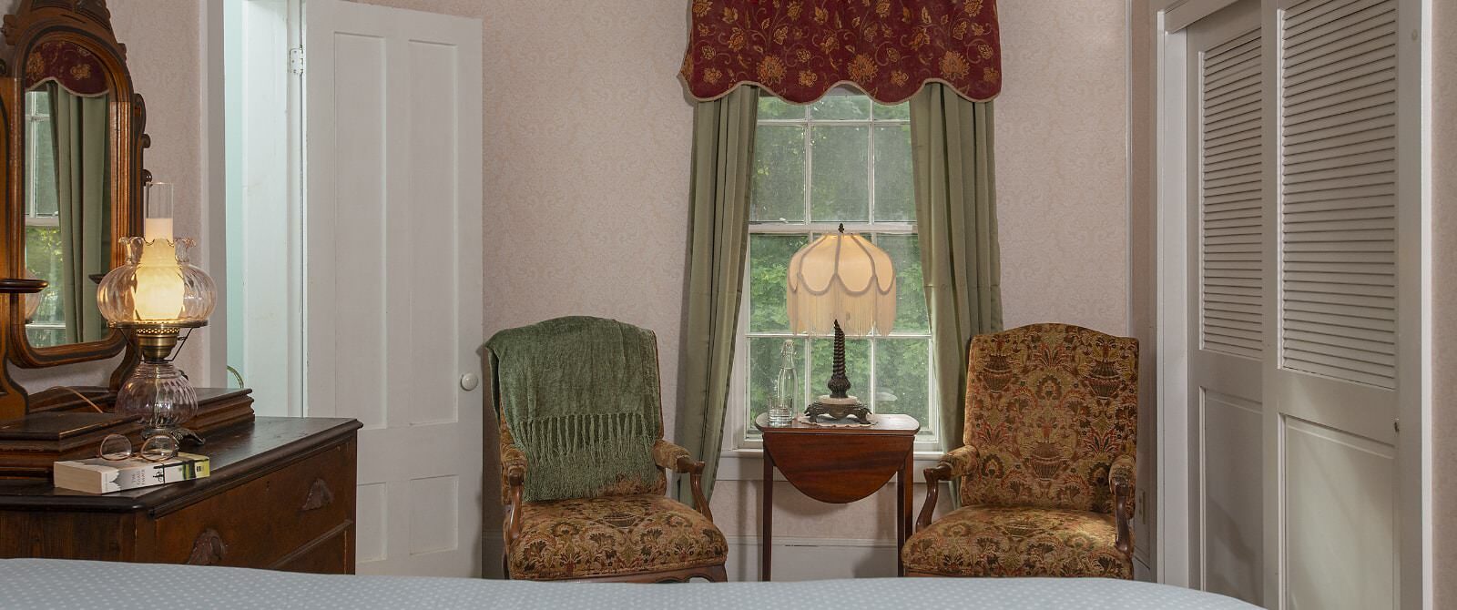 Bedroom with light pink brocade wallpaper, antique wooden dresser, light blue bedding, sitting area, and window with a view of green trees