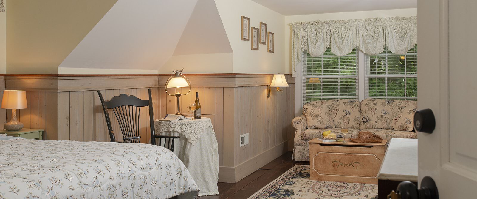 Bedroom with white walls, light wooden wainscoting, floral bedding, sitting area, floral upholstered loveseat, and wooden trunk
