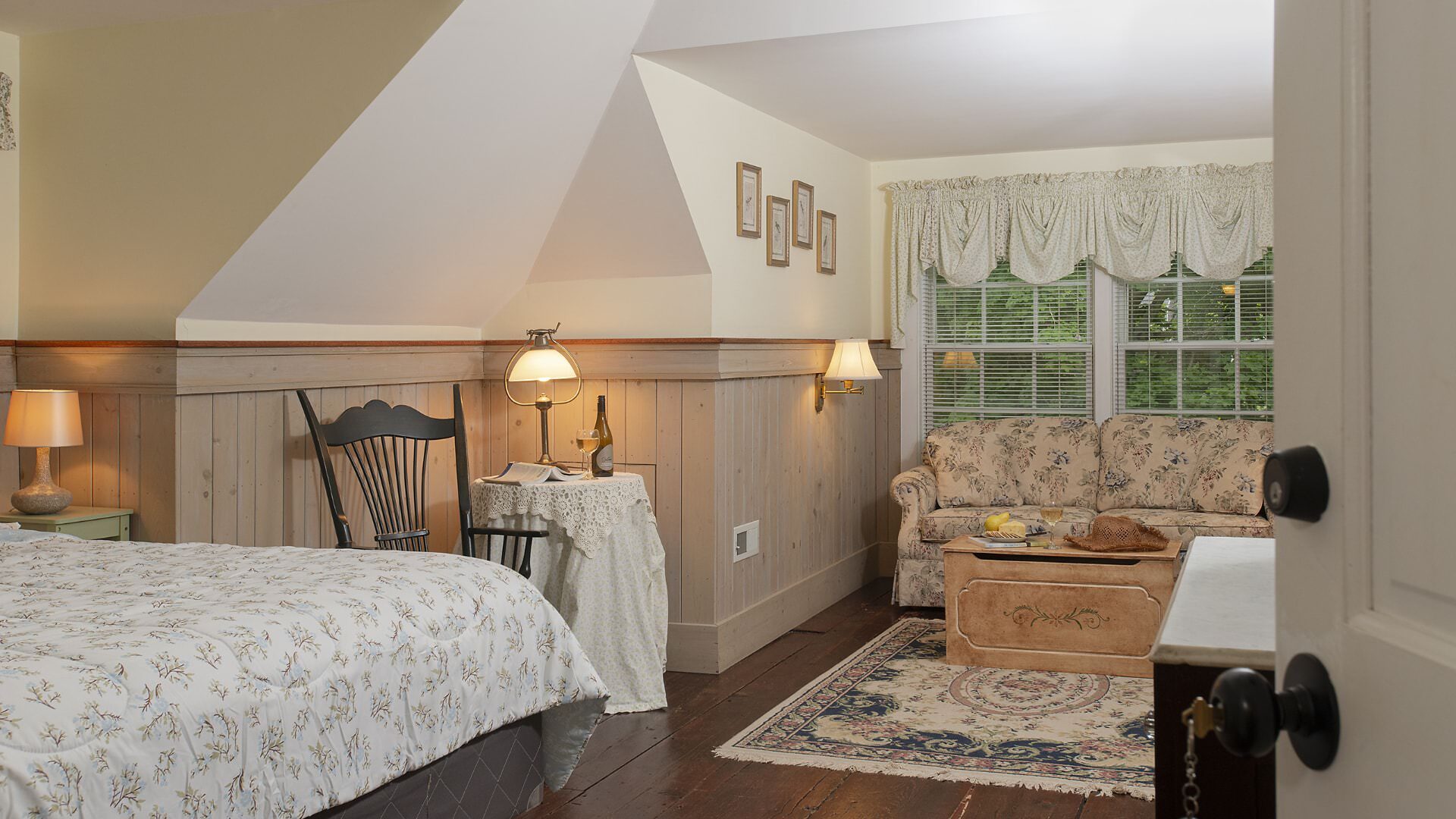 Bedroom with white walls, light wooden wainscoting, floral bedding, sitting area, floral upholstered loveseat, and wooden trunk