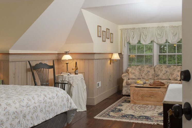 Bedroom with white walls, light wooden wainscoting, floral bedding, sitting area, floral upholstered loveseat, and wooden trunk