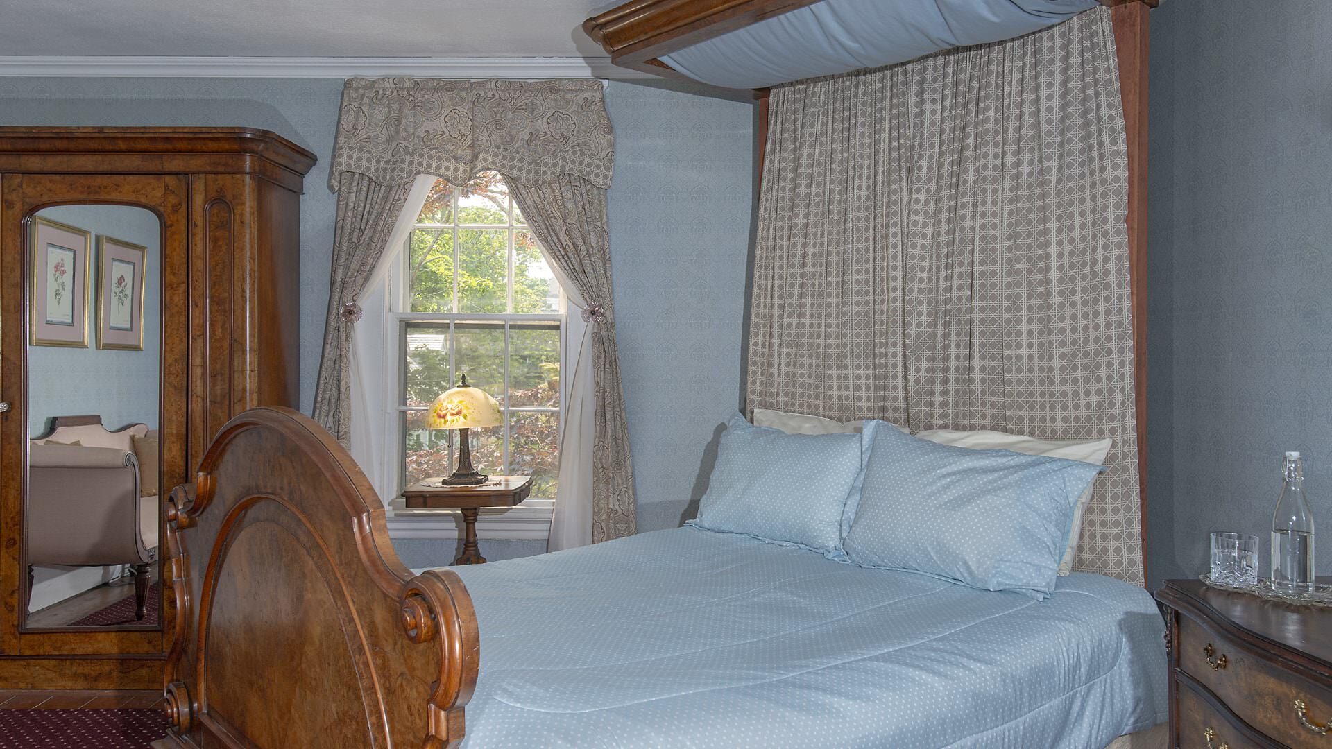 Bedroom with light blue wallpaper, carpeting, antique canopy bed, light blue bedding, and antique dark wooden hutch and dresser