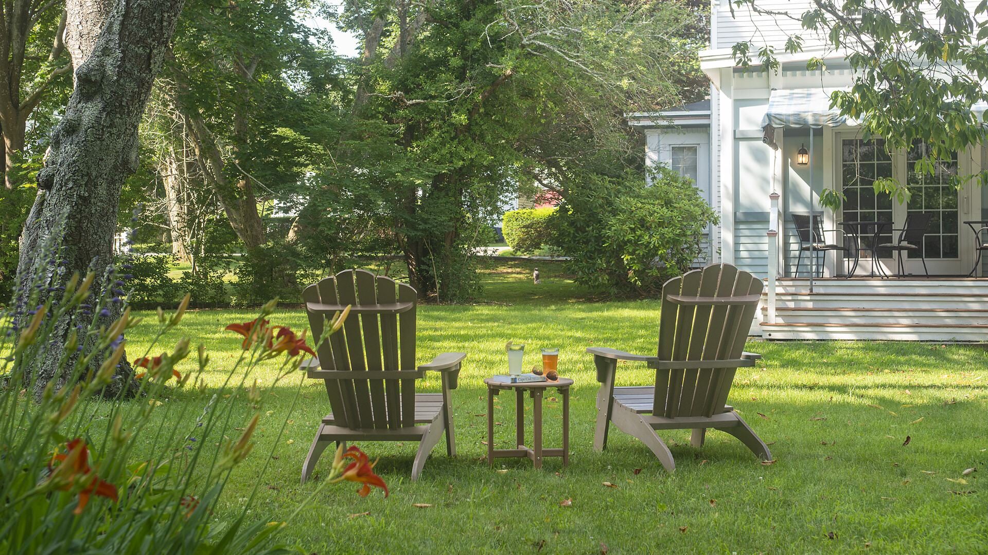 Exterior view of property painted light blue with white trim surrounded by large green trees and green yard with two Adirondack chairs