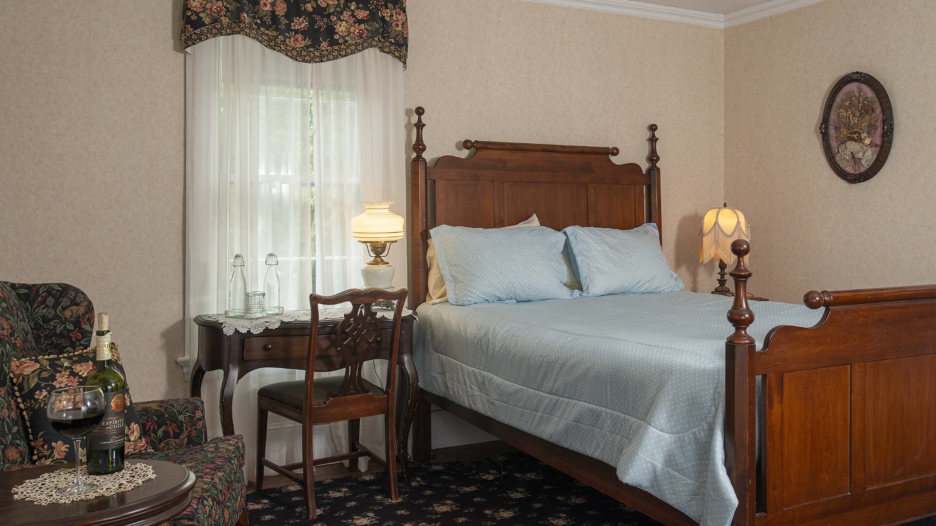 Bedroom with light peach wallpaper, antique wooden headboard and footboard, light blue bedding, sitting area, and antique wooden desk with chair