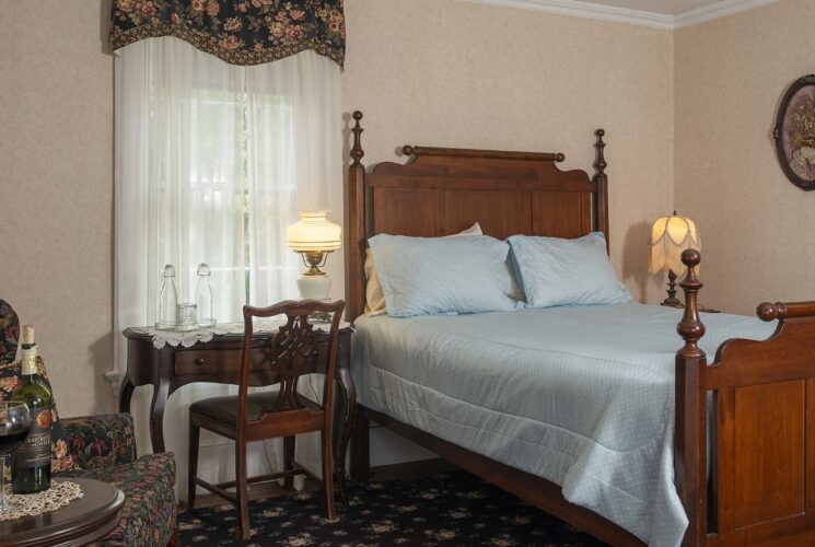 Bedroom with light peach wallpaper, antique wooden headboard and footboard, light blue bedding, sitting area, and antique wooden desk with chair
