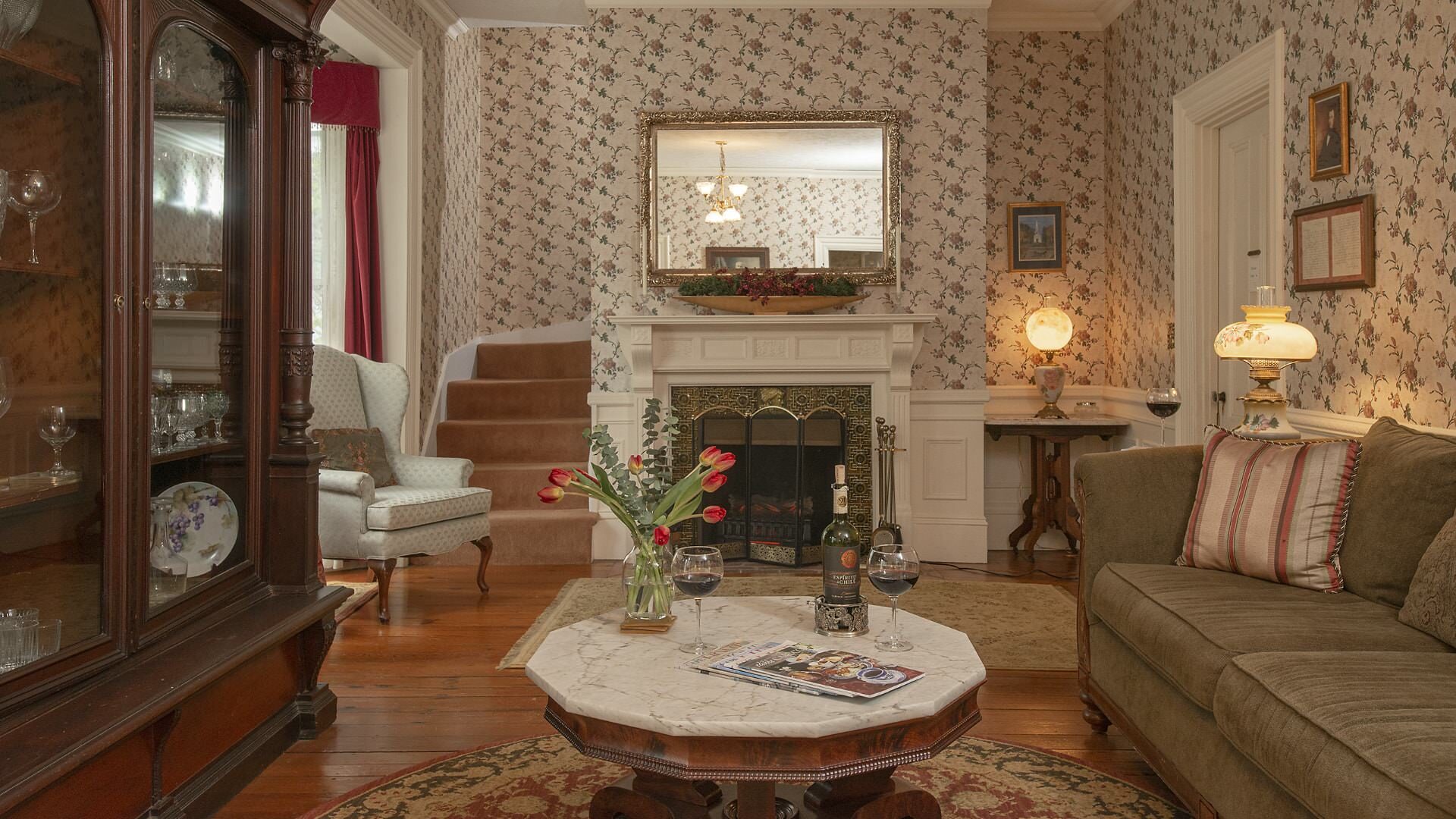 Living room with floral wallpaper, hardwood flooring, antique china cabinet, tan upholstered couch, and antique upholstered armchairs