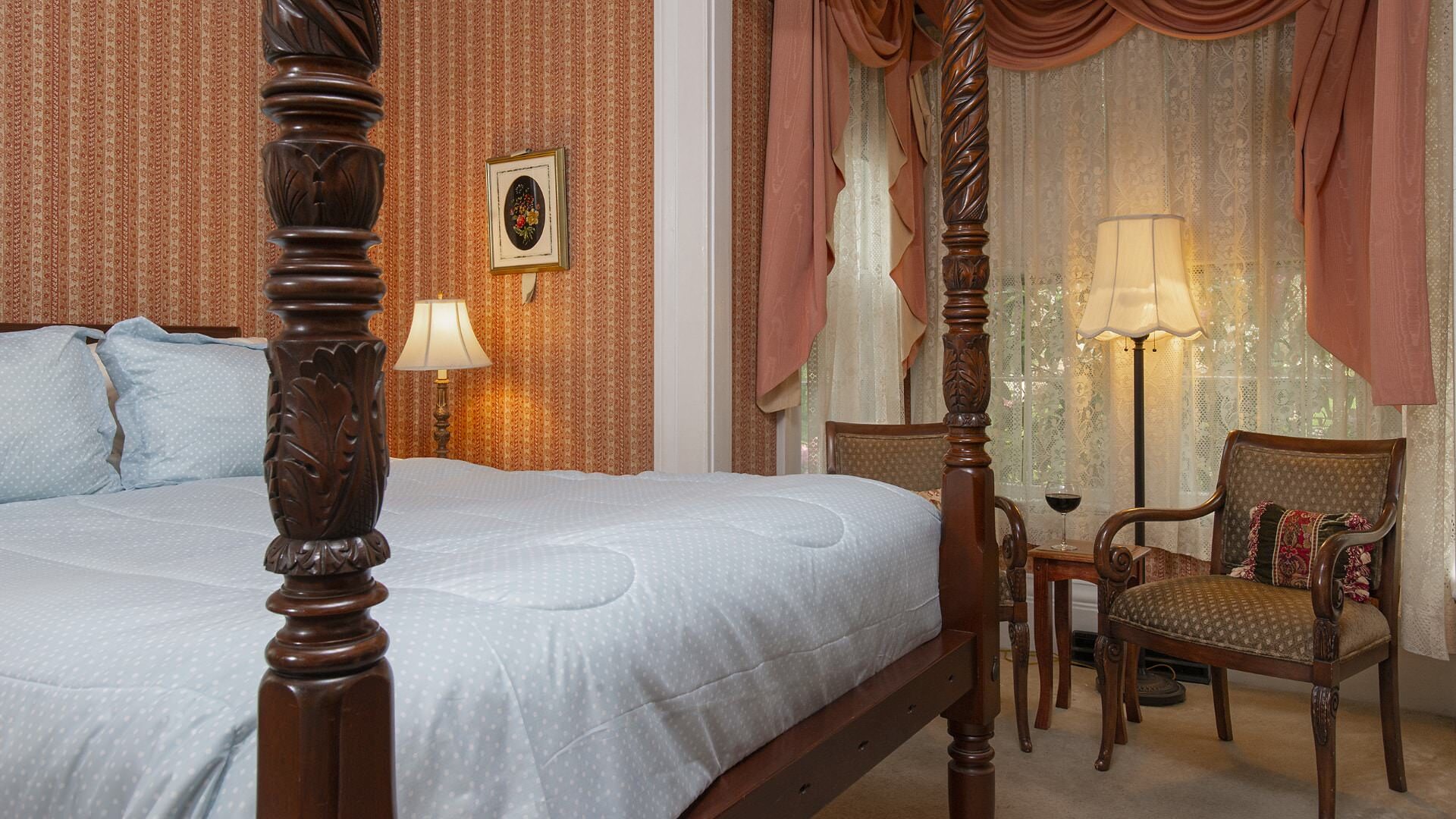 Bedroom with light colored striped wallpaper, carpeting, dark wooden four-poster bed, light blue bedding, and sitting area