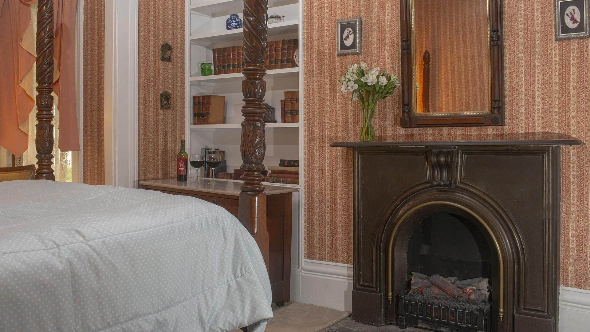 Bedroom with striped looking wallpaper, white trim, carpeting, four-poster wooden bed, and antique fireplace