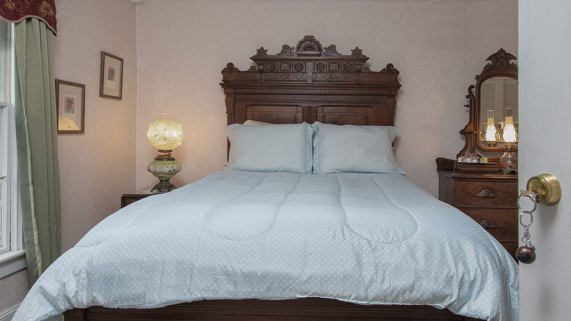 Bedroom with light pink brocade wallpaper, antique wooden headboard, light blue bedding, and dark wooden dresser with mirror