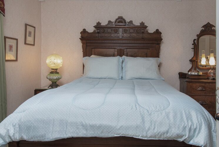 Bedroom with light pink brocade wallpaper, antique wooden headboard, light blue bedding, and dark wooden dresser with mirror