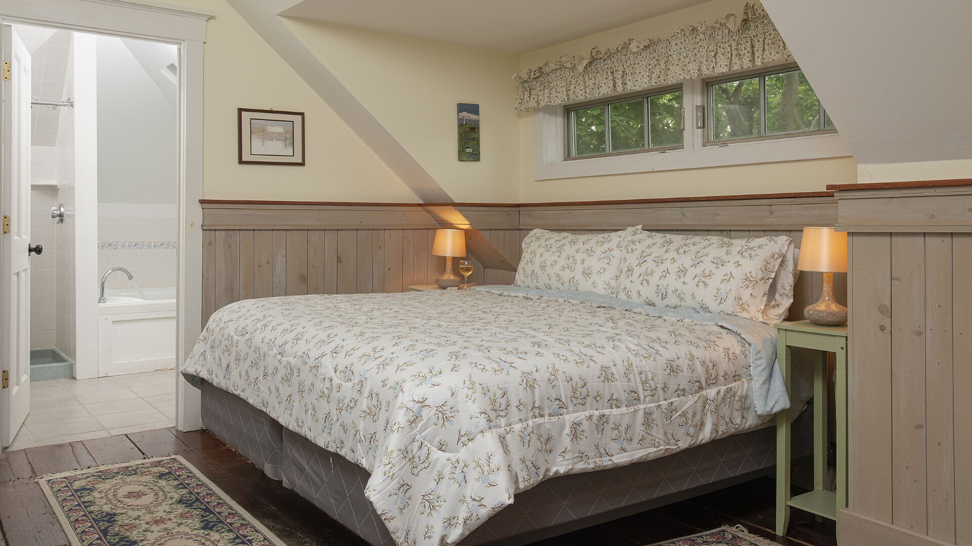 Bedroom with white walls, light wooden wainscoting, floral bedding, and view into bathroom