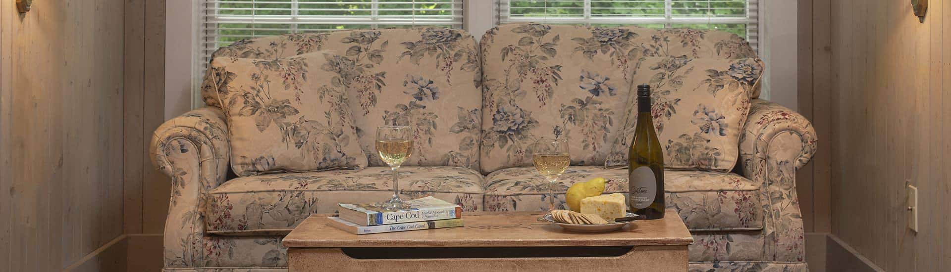 Floral upholstered loveseat and coffee table with Cape Cod books, wine filled glasses, and plate with cheese, crackers, and a pear