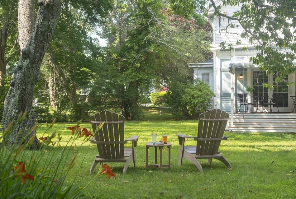 Exterior view of property painted light blue with white trim surrounded by large green trees and green yard with two Adirondack chairs