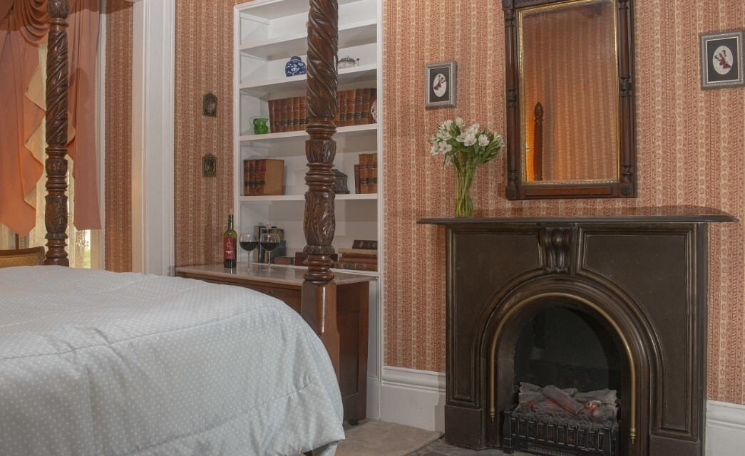 Bedroom with striped looking wallpaper, white trim, carpeting, four-poster wooden bed, and antique fireplace