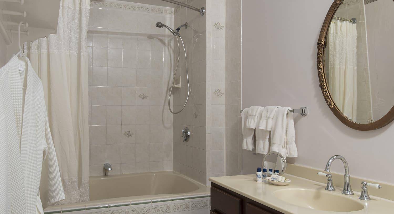 Bathroom with light walls, light tiled bathtub with shower, dark wooden vanity with cream counter, and oval framed mirror