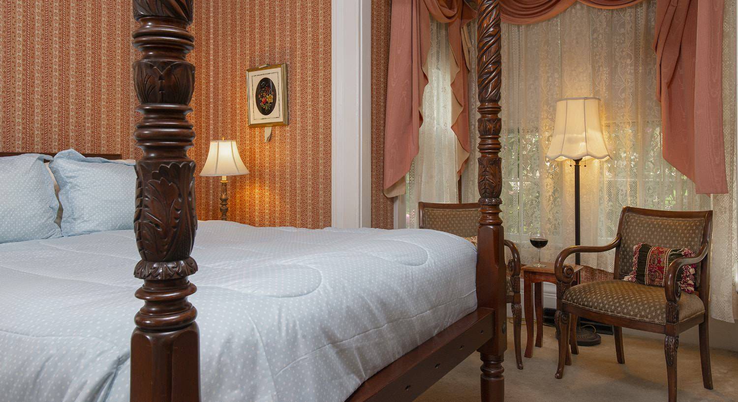 Bedroom with light colored striped wallpaper, carpeting, dark wooden four-poster bed, light blue bedding, and sitting area
