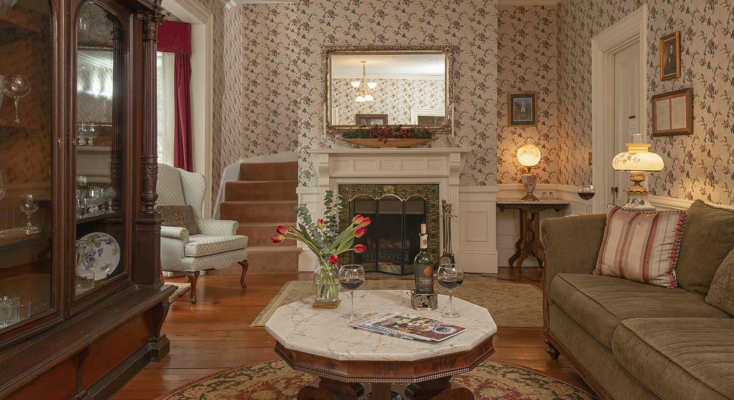 Living room with floral wallpaper, hardwood flooring, antique china cabinet, tan upholstered couch, and antique upholstered armchairs