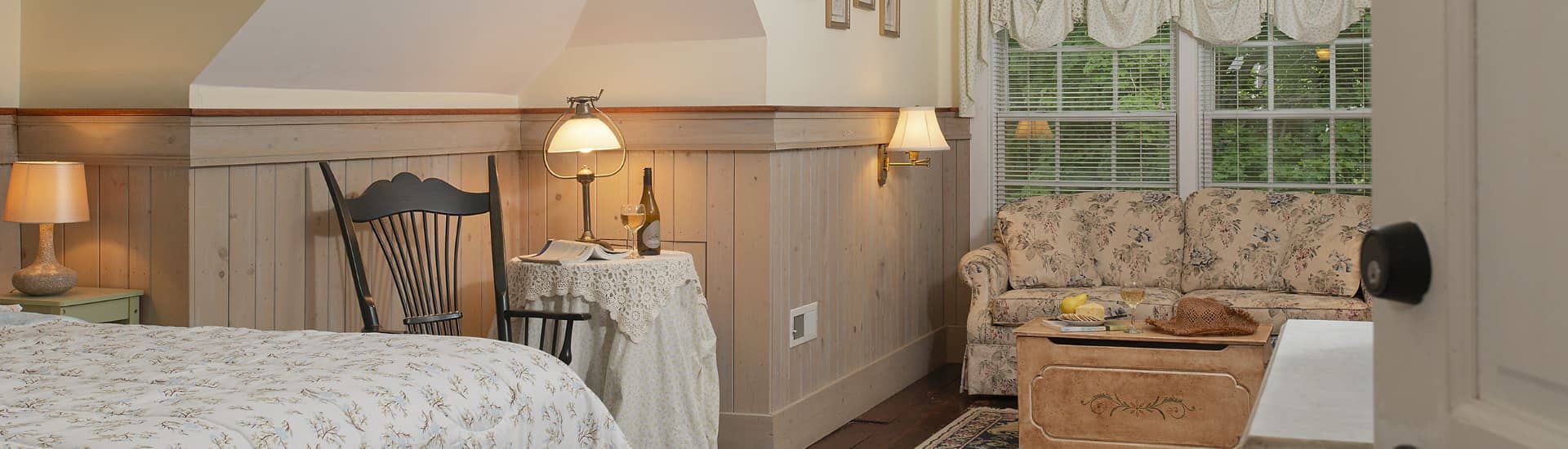 Bedroom with white walls, light wooden wainscoting, floral bedding, sitting area, floral upholstered loveseat, and wooden trunk